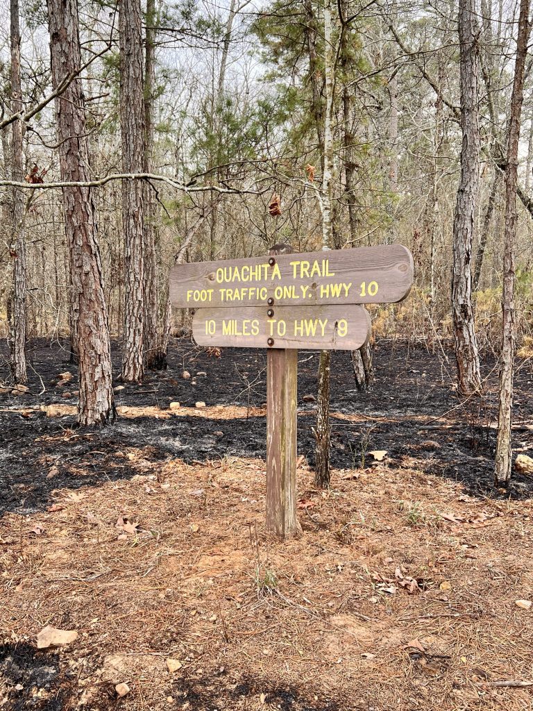 A wooden trail sign marking the Ouachita Trail, indicating foot traffic only, with directions to Highway 10 and a distance of 10 miles to Highway 9, surrounded by trees and burned ground.