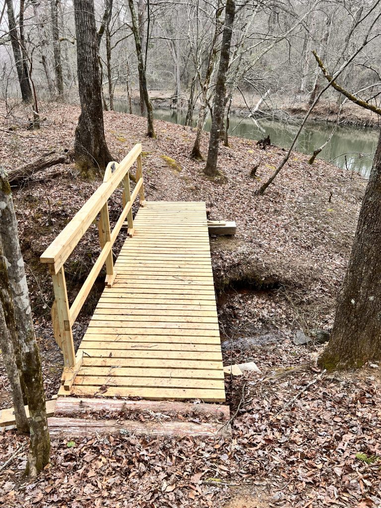 A wooden bridge spans a small creek, surrounded by bare trees and fallen leaves in a forested area.