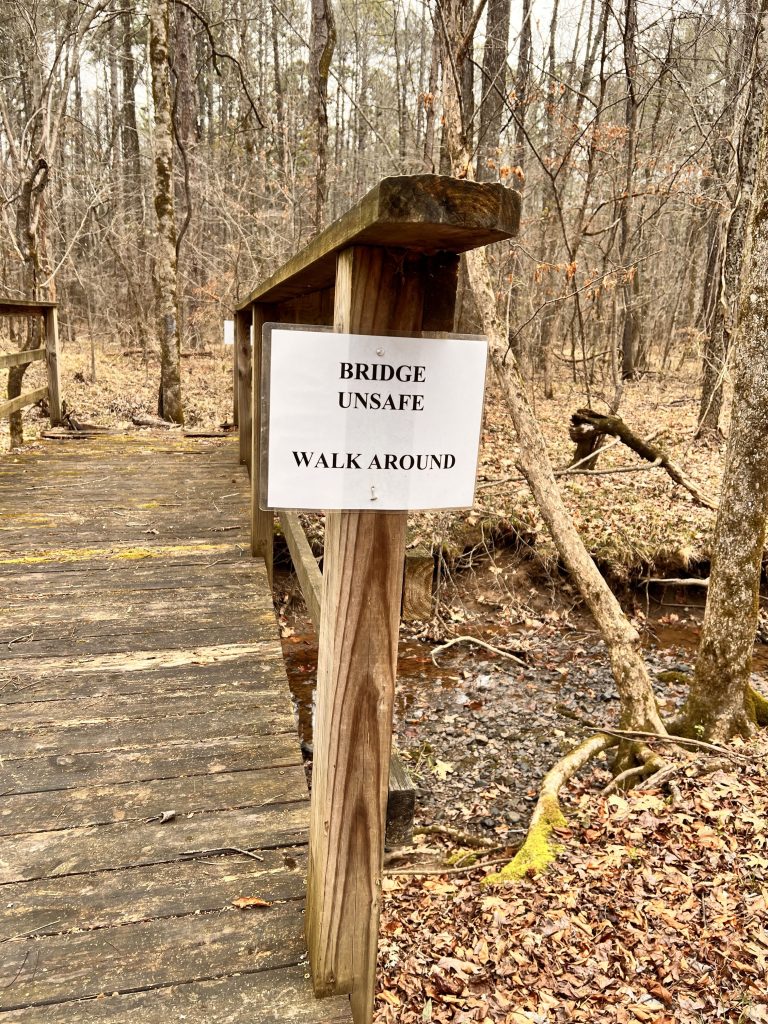 A wooden bridge with a sign reading 'BRIDGE UNSAFE WALK AROUND,' set in a wooded area with fallen leaves and a small creek.