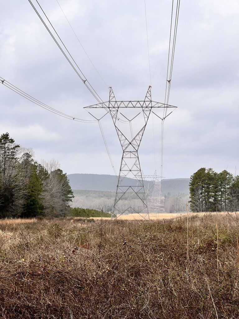 A power line tower stands in a grassy field with cloudy skies in the background, surrounded by trees and rolling hills.
