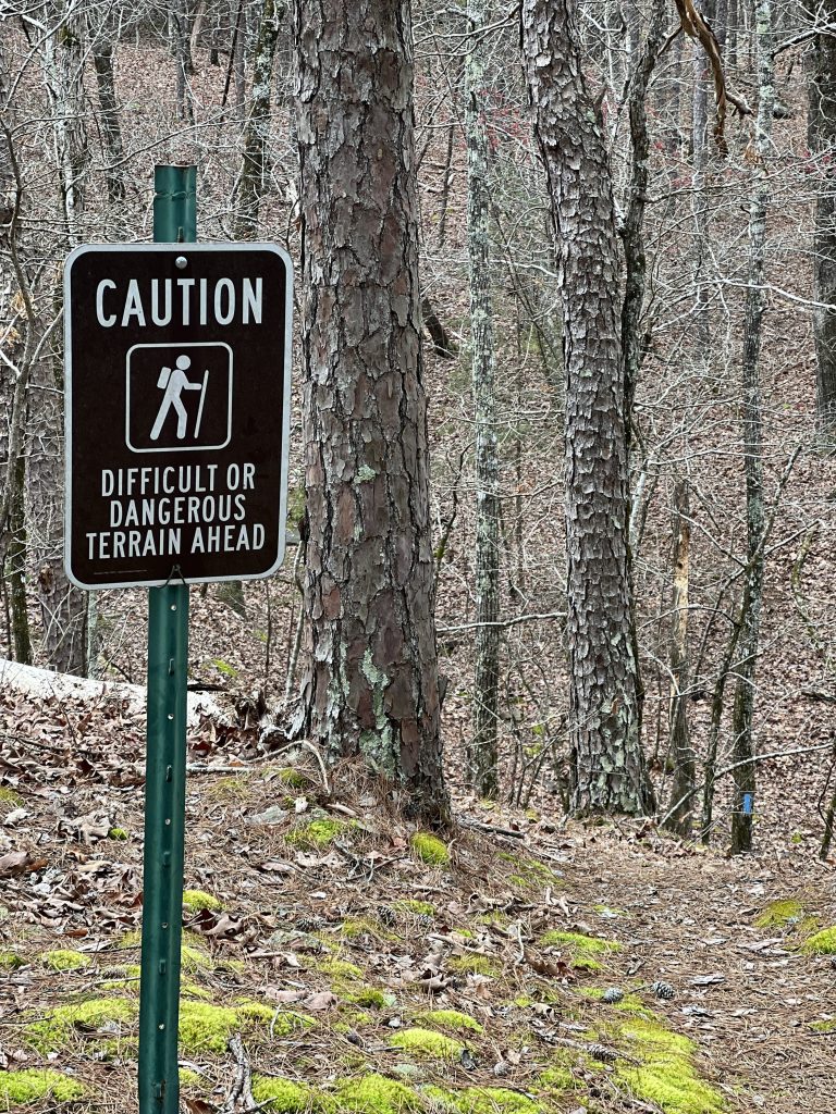 A caution sign warning of difficult or dangerous terrain ahead, with trees and forest floor visible in the background.