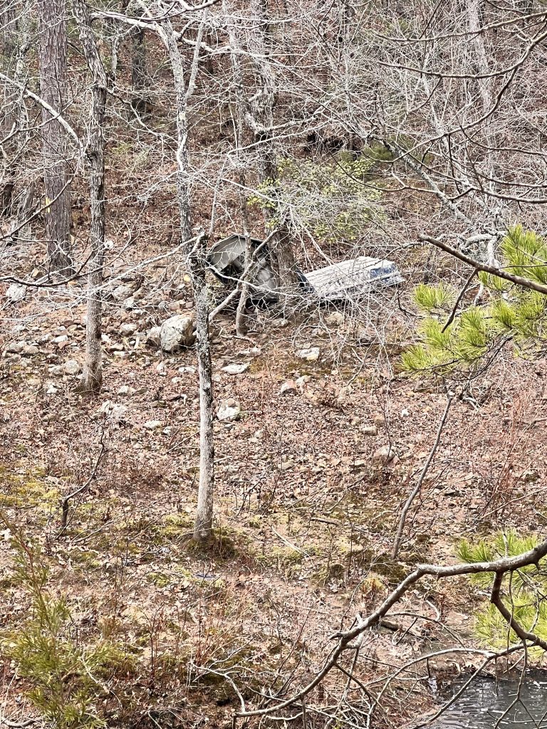 A partially obscured abandoned vehicle in a dry, rocky forested area with sparse trees and bare branches.