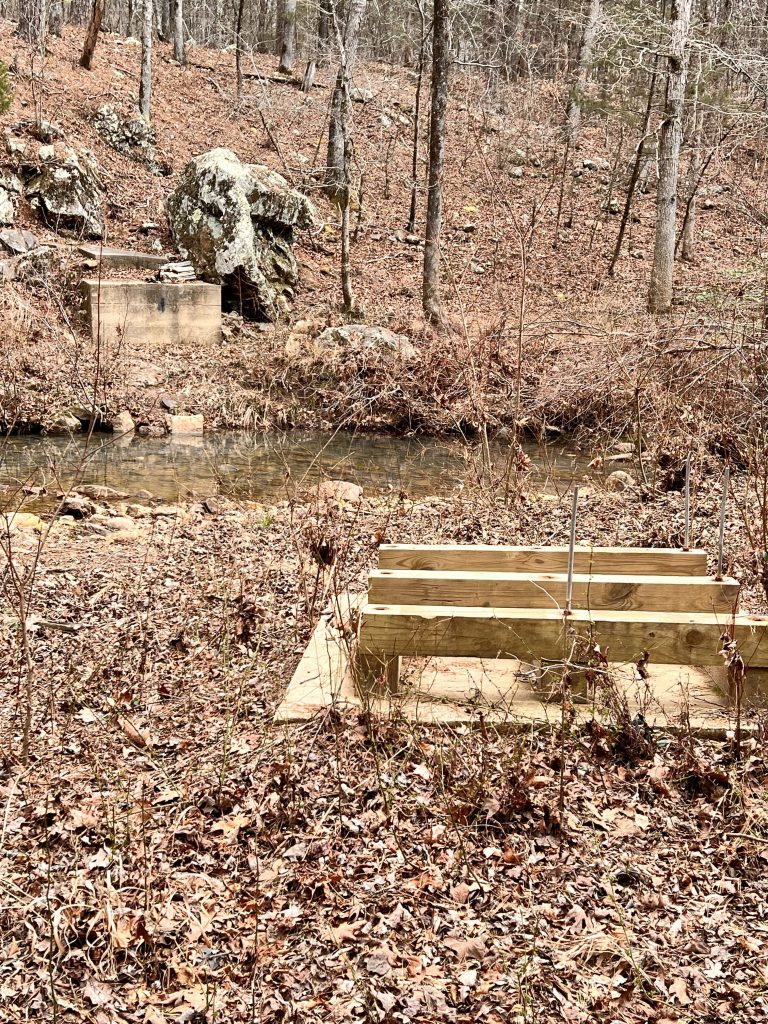 A wooden platform sits in a semi-dry area next to a shallow creek, surrounded by bare trees and rocky terrain in a forested setting.