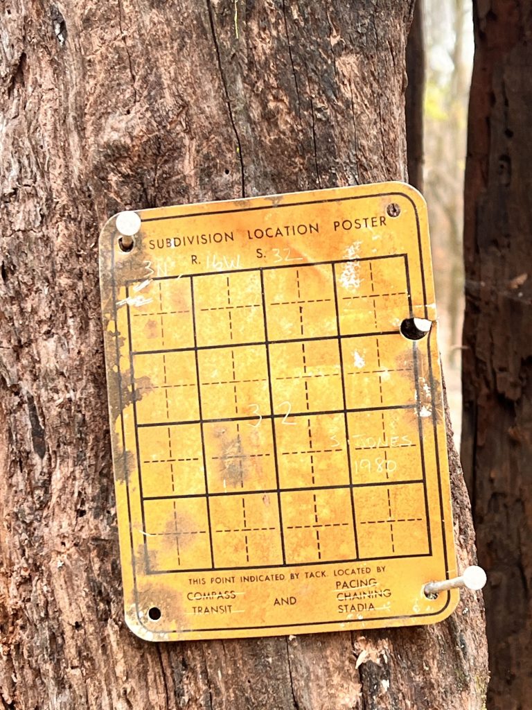 A weathered yellow subdivision location poster attached to a tree, featuring a grid pattern with markings, dated 1980. The poster includes instructions for locating the point using compass transit and pacing.